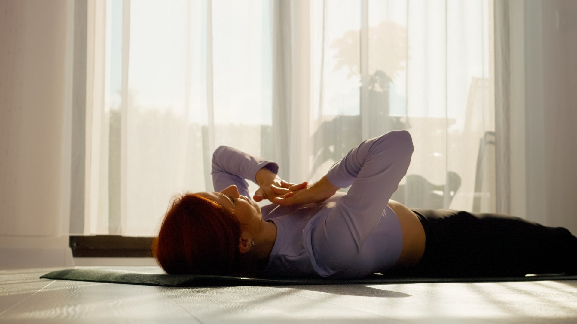 In a bright living room, a woman engages in stretching and pilates exercises on a mat. The sunny space promotes a healthy morning routine filled with movement.